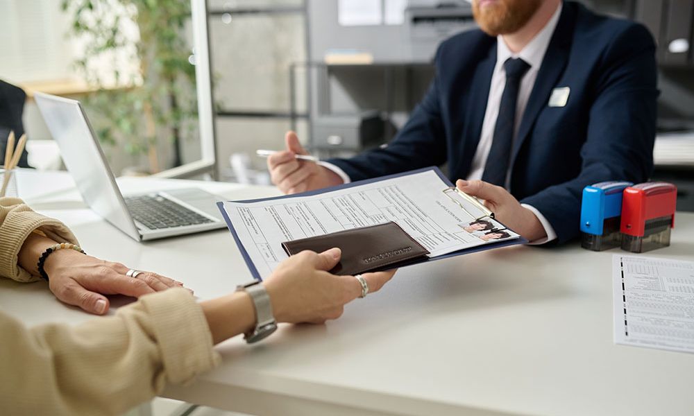 Person handing over documents to a business professional across a desk with a laptop, stamps, and papers.