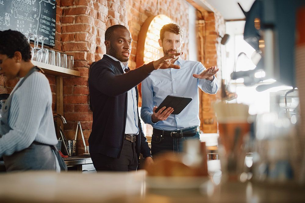Two men discussing work with a tablet in a trendy cafe, while a barista prepares a drink in the background.