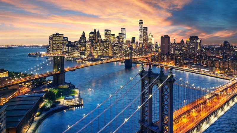 Aerial view of New York City skyline at sunset with lit bridges and skyscrapers reflecting in the river.