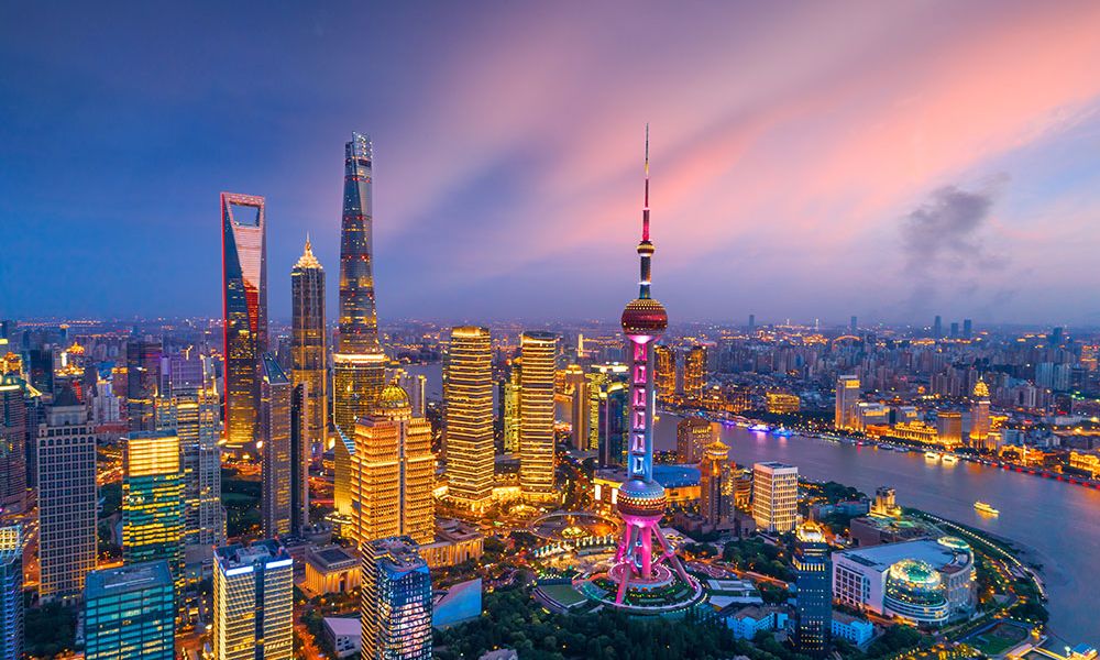 Shanghai cityscape at sunset with lit skyscrapers and the iconic Oriental Pearl Tower reflecting on the Huangpu River.