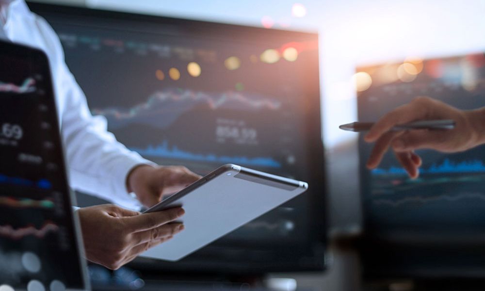 Business team analyzing financial data on tablet and screens, stock market graphs visible in background.