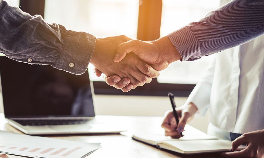 Business handshake in office, agreement concept, two people shaking hands near a laptop and documents on a desk.