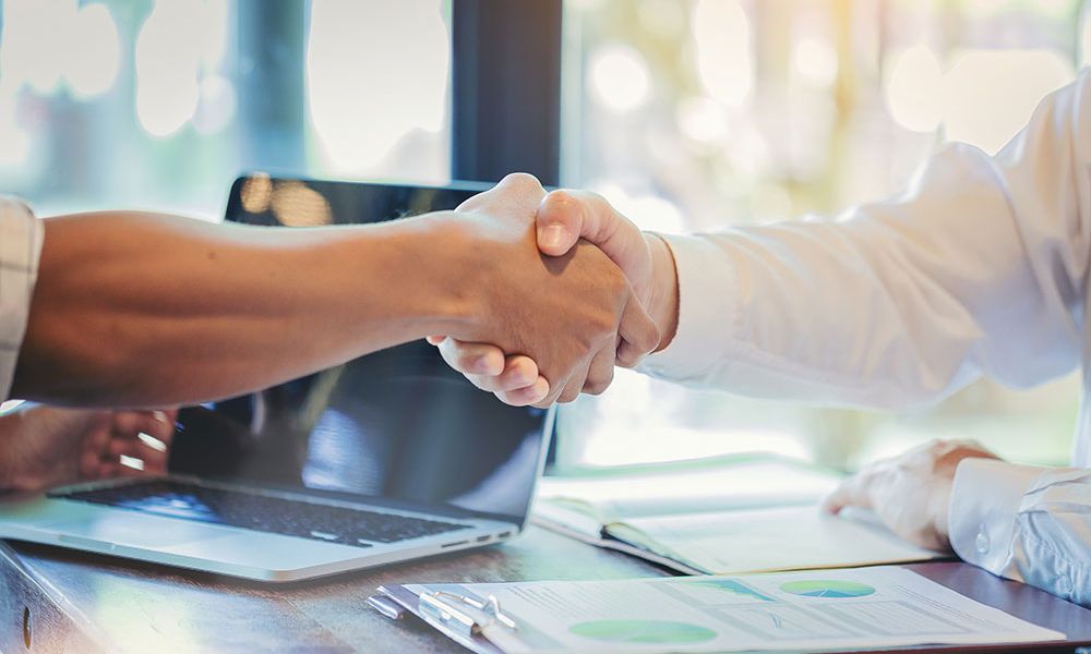 Handshake over a desk with a laptop and documents, symbolizing a successful business partnership.