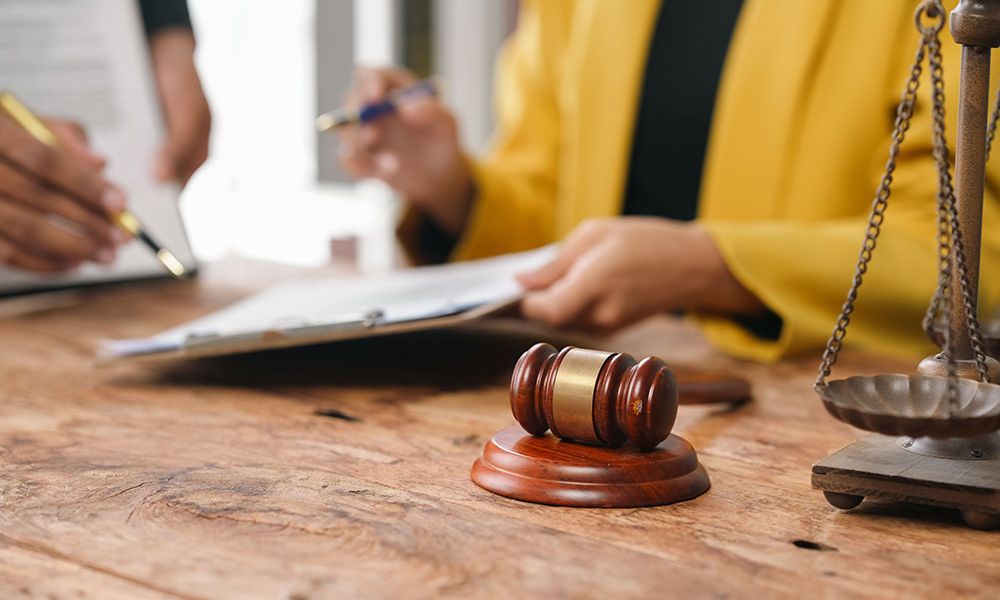 Legal consultation scene with gavel on table, people reviewing documents, and scales of justice in the background.