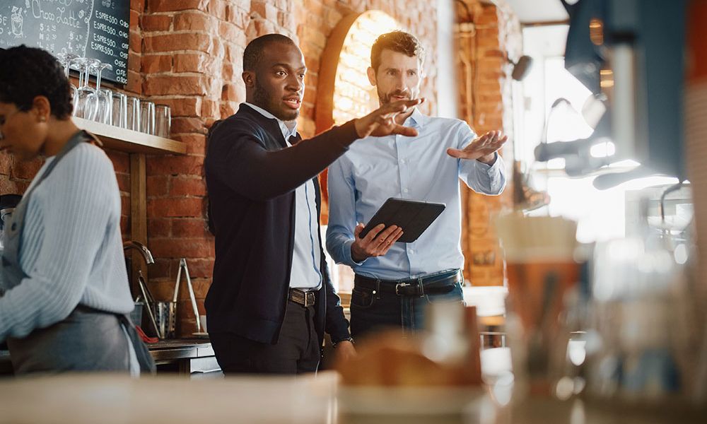 Two men discussing work with a tablet in a trendy cafe, while a barista prepares a drink in the background.