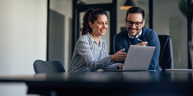 Two colleagues smiling while working on a laptop in a modern office setting.