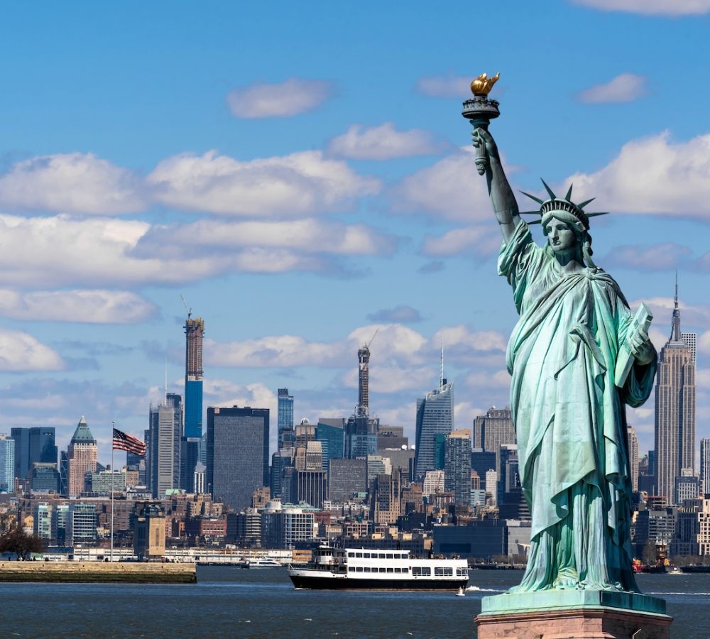 Statue of Liberty with New York City skyline in the background under a blue sky with clouds.