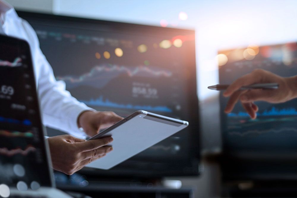 Business team analyzing financial data on tablet and screens, stock market graphs visible in background.