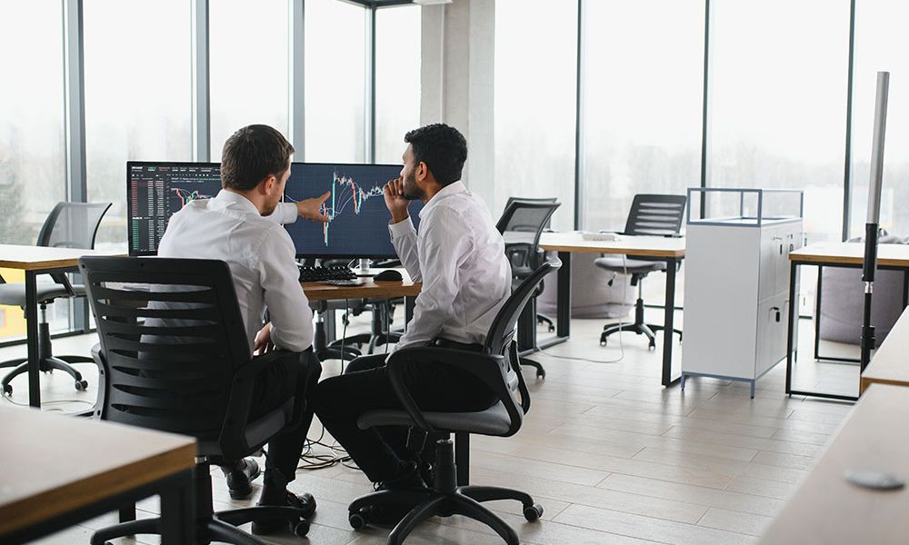 Two businessmen analyzing financial graphs on a computer in a modern office setting.