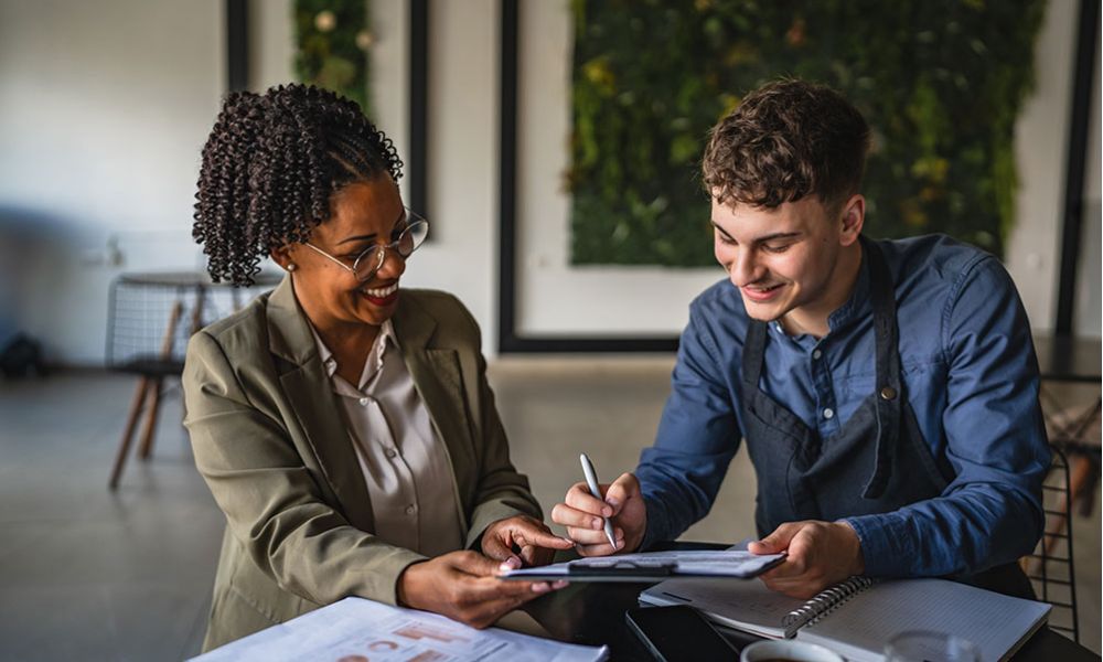 Two colleagues happily collaborating at a desk with documents and coffee in an office setting.
