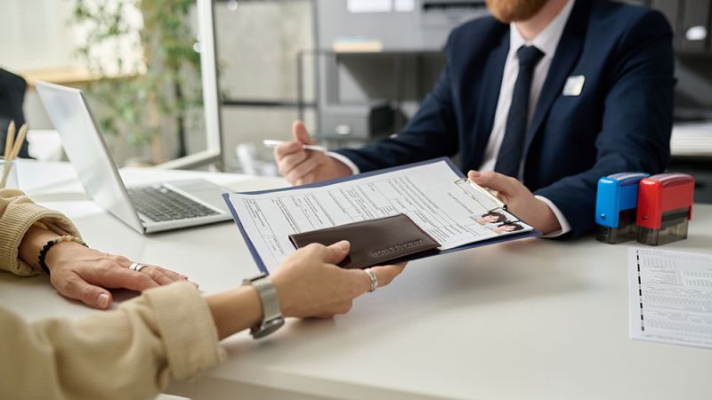 Person handing over documents to a business professional across a desk with a laptop, stamps, and papers.