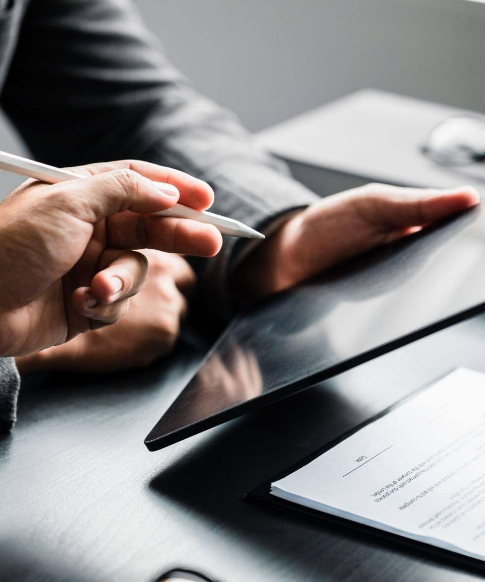 Hands hold a tablet while another hand points with a stylus. A document with a pen lies on a dark table, near a pair of glasses in a professional setting.