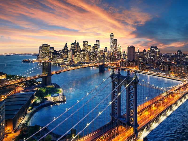 Aerial view of New York City skyline at sunset with lit bridges and skyscrapers reflecting in the river.