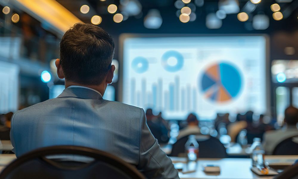 Man in blue suit at a conference, looking at a screen displaying colorful charts and graphs, business presentation setting.