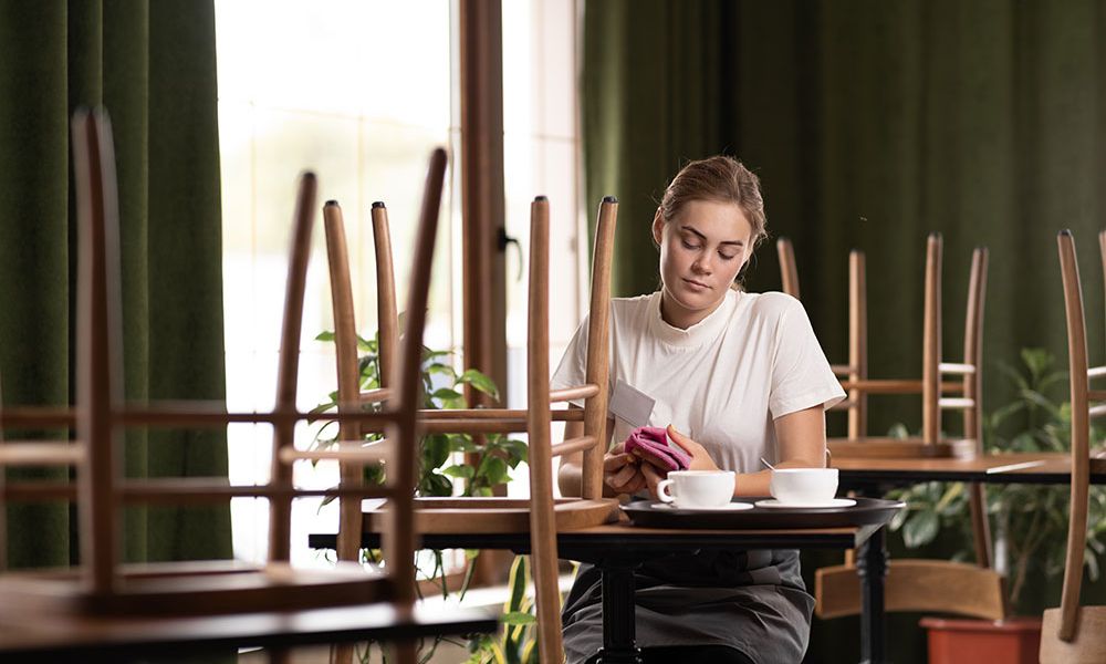 Woman seated in empty café with stacked chairs, using phone and drinking coffee.