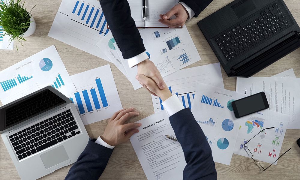 Two businesspeople shaking hands over a contract and financial charts on a desk with laptops and a smartphone.