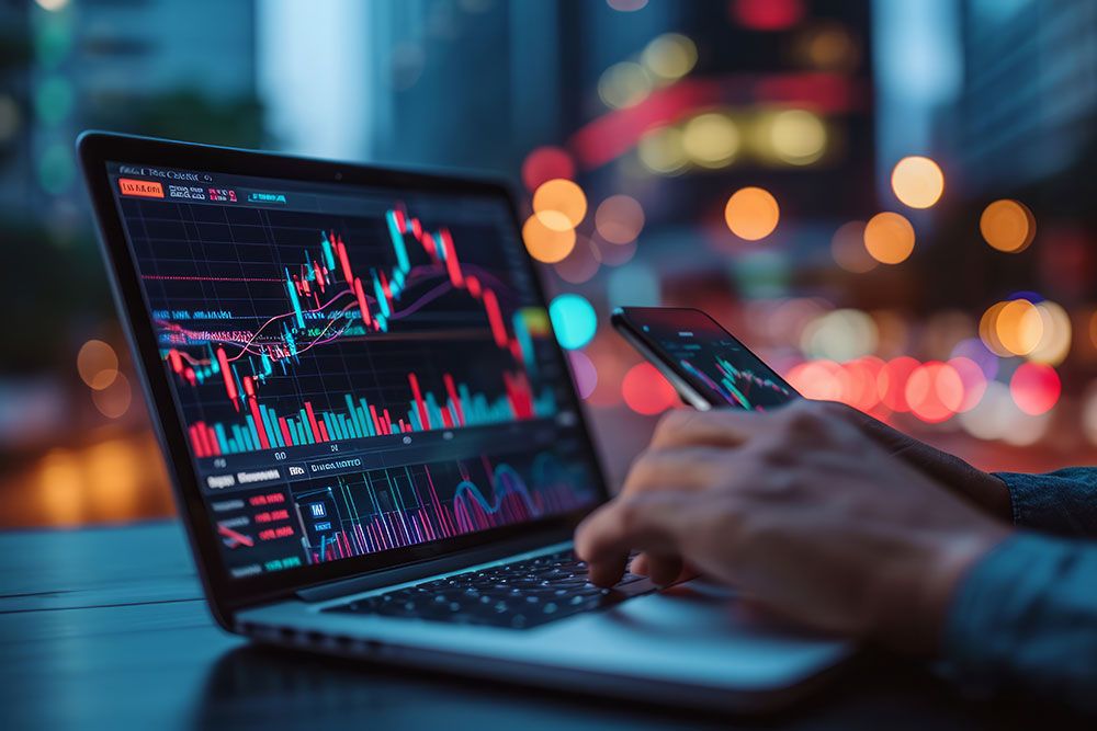 Person analyzing stock market data on a laptop and smartphone in a cityscape setting at night.