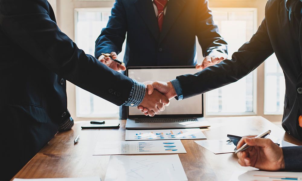 Business professionals shaking hands over a meeting table with documents and a laptop, symbolizing agreement and partnership.