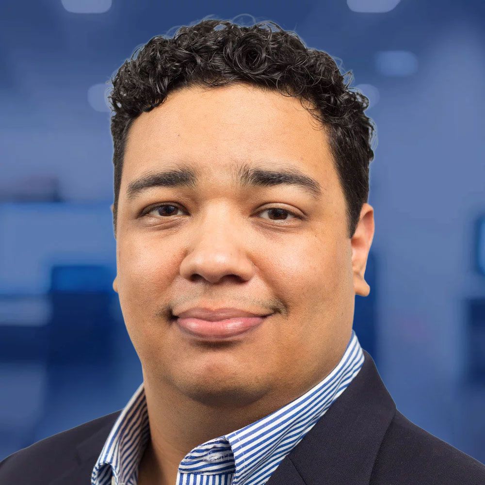 Professional man in suit with curly hair smiles in a corporate setting, blurred office background.
