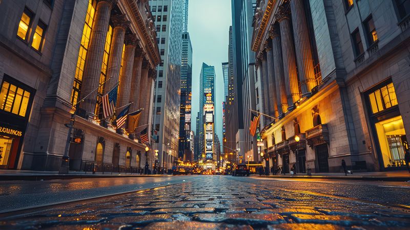 Downtown city street with illuminated buildings and wet cobblestones, flags hanging from columns, and a fading skyline.