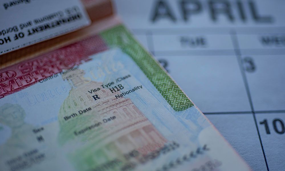 Close-up of an H-1B visa on a passport with a blurred calendar showing the month of April in the background.