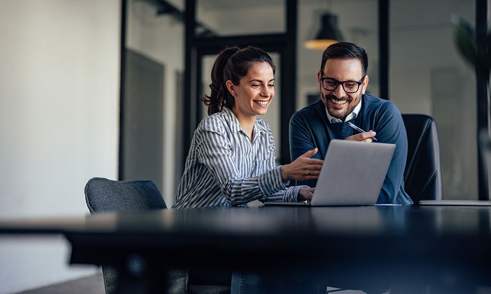 Two colleagues smiling while working on a laptop in a modern office setting.