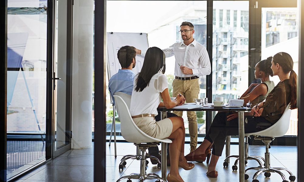 Business meeting presentation with diverse team in a bright office, featuring a man pointing at a whiteboard.