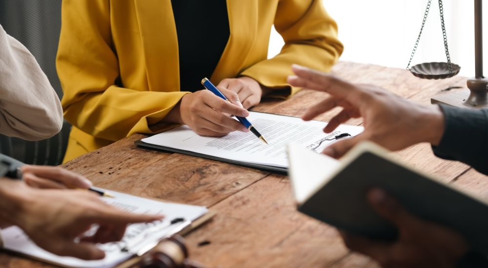 People discussing and signing legal documents at a wooden table, featuring a gavel and scale of justice.