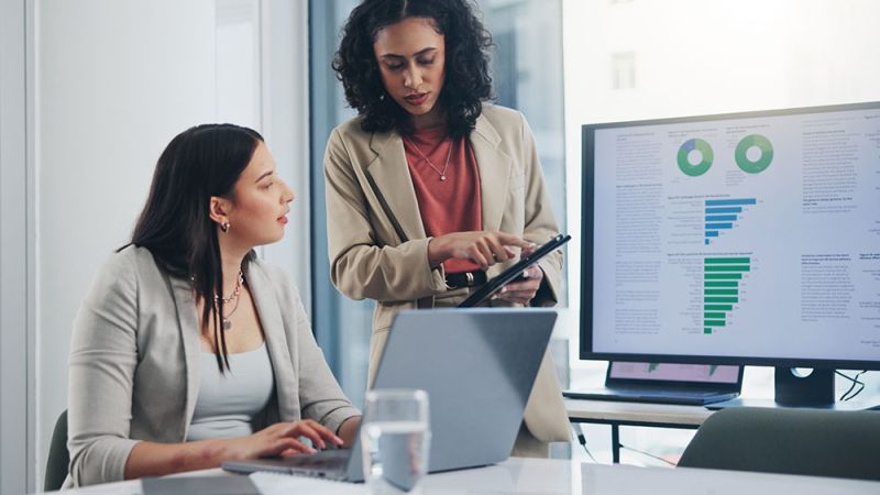 Two businesswomen discussing data on a tablet, with charts displayed on a computer screen in a modern office setting.