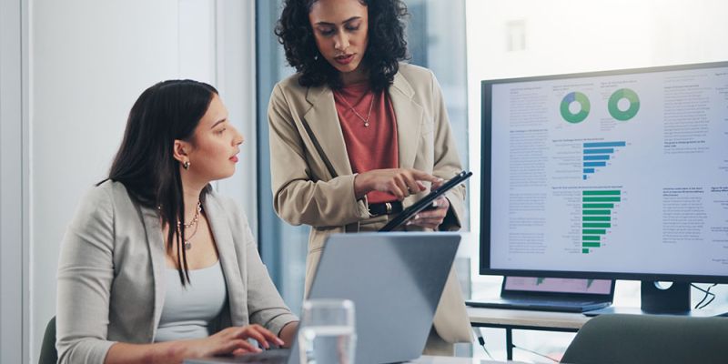 Two businesswomen discussing data on a tablet, with charts displayed on a computer screen in a modern office setting.