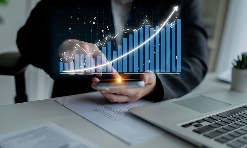 Person analyzes digital financial chart with growth trends on a smartphone, sitting at a desk with a laptop.