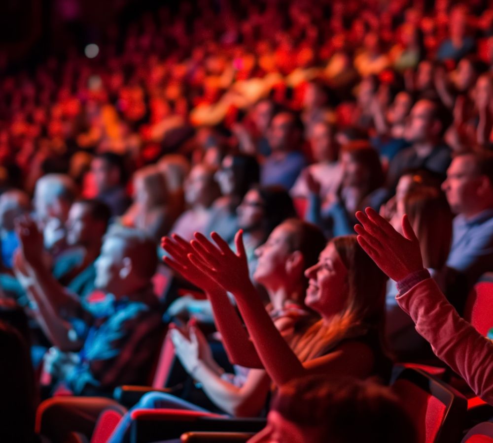 Audience clapping in a packed theater, vibrant red lighting, engaging performance atmosphere.