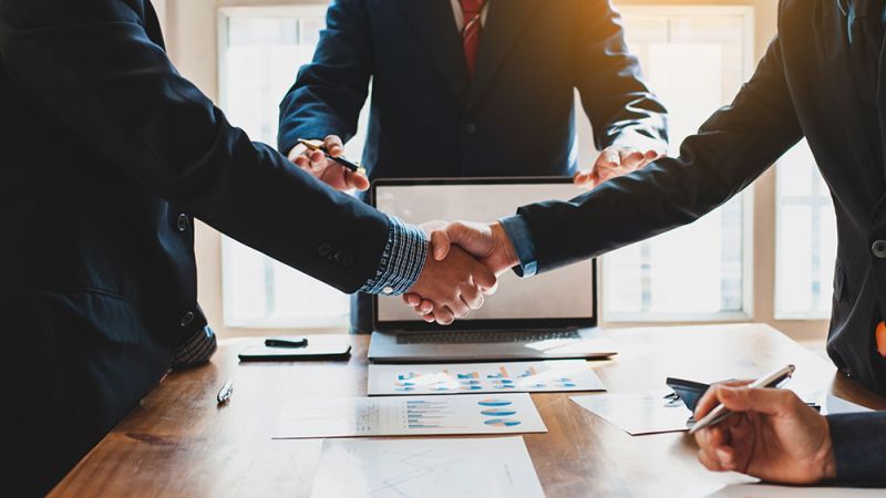 Business professionals shaking hands over a meeting table with documents and a laptop, symbolizing agreement and partnership.