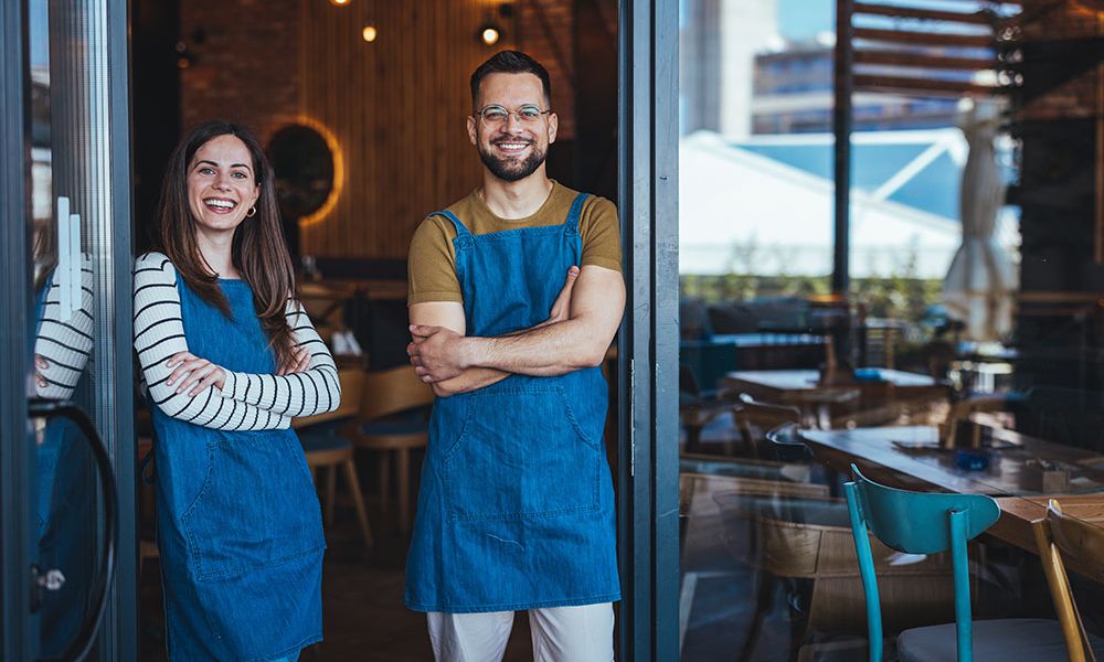 Two smiling cafe owners in blue aprons stand at the entrance of their cozy, modern coffee shop.